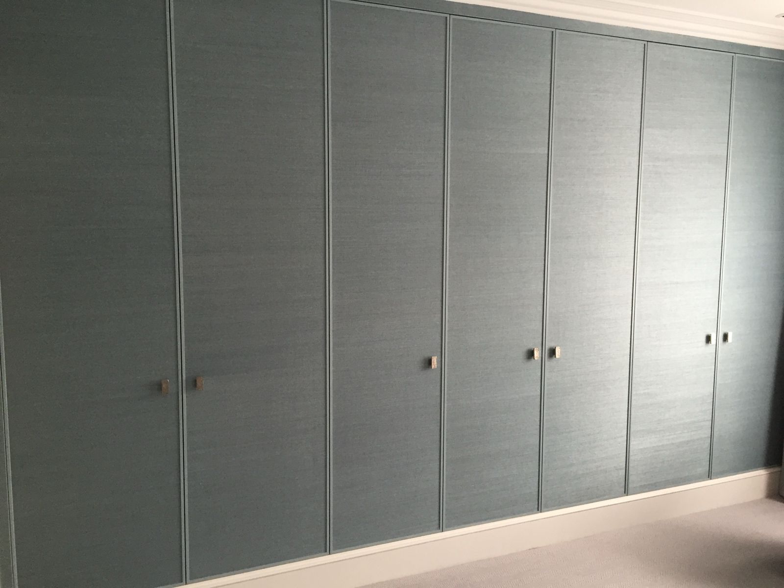 Wall of fitted wardrobes faced in blue grasscloth with bronze pulls